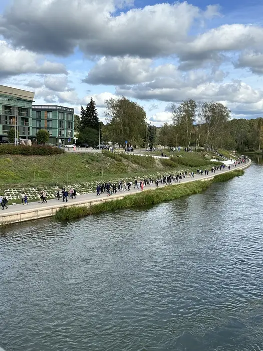 High-angle view of the Neris River in Vilnius with a paved walking path on the bank, where many people are walking past modern green buildings and grassy slopes under a cloudy blue sky.