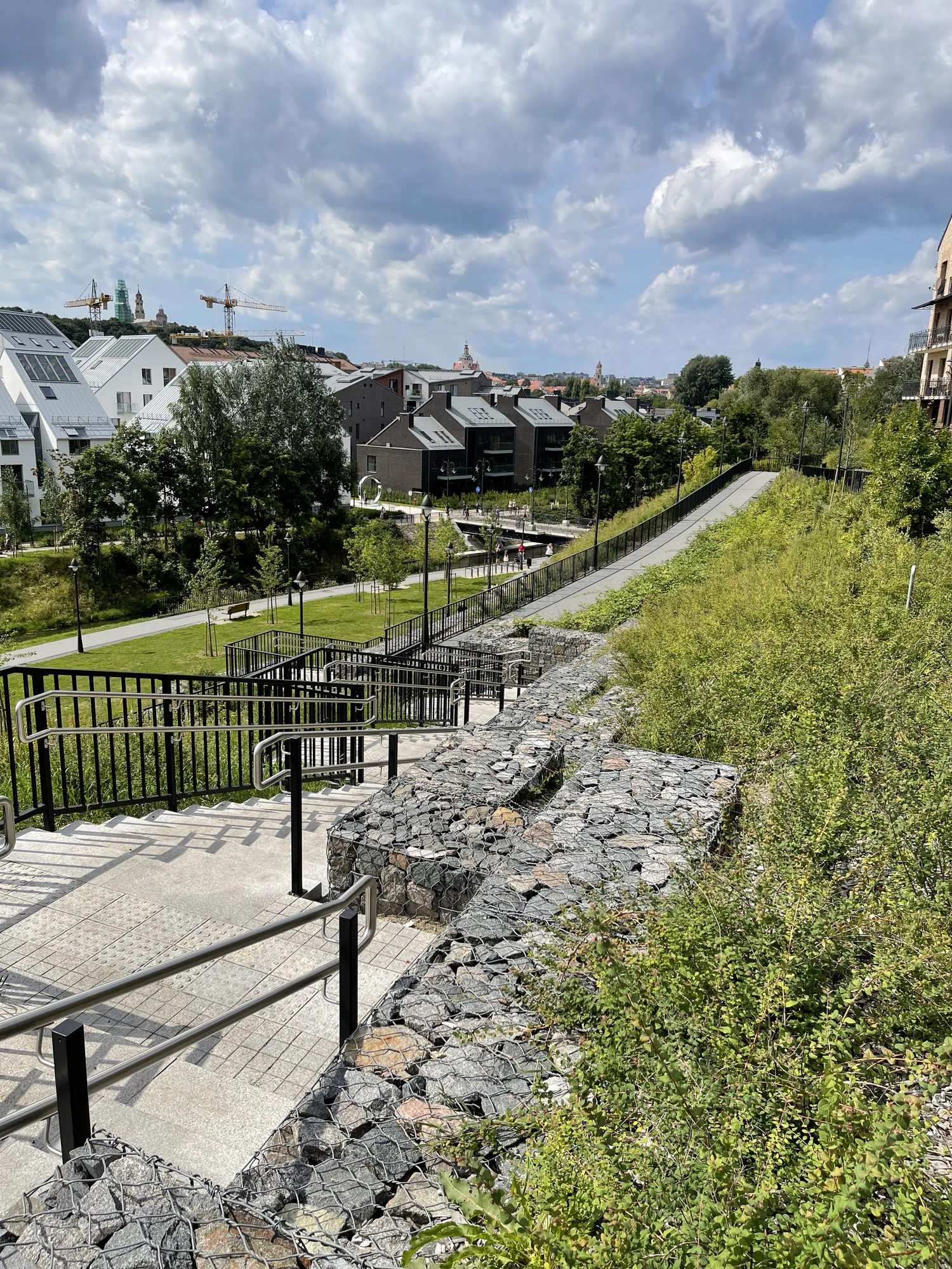 A high-angle view of a modern paved pedestrian path and stairs with metal handrails winding down a grassy hill lined with gabion stone walls, overlooking contemporary residential buildings in Vilnius.