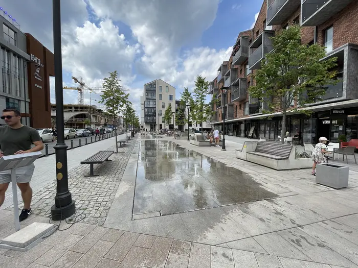 A wide-angle view of a modern pedestrian street in the Paupys district of Vilnius, featuring a central flat water fountain, modern brick apartment buildings with cafes, young trees, and people relaxing on benches.