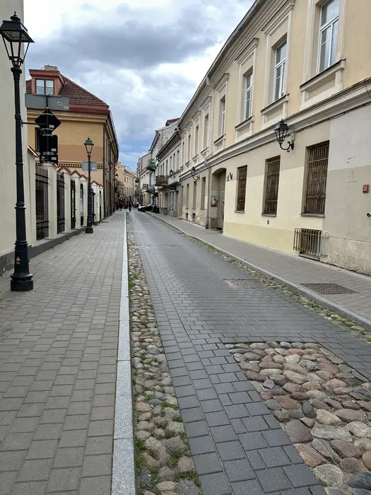 A narrow street in Vilnius Old Town with grey paving stones, cobblestone sections, historic buildings, and vintage street lamps under a cloudy sky.