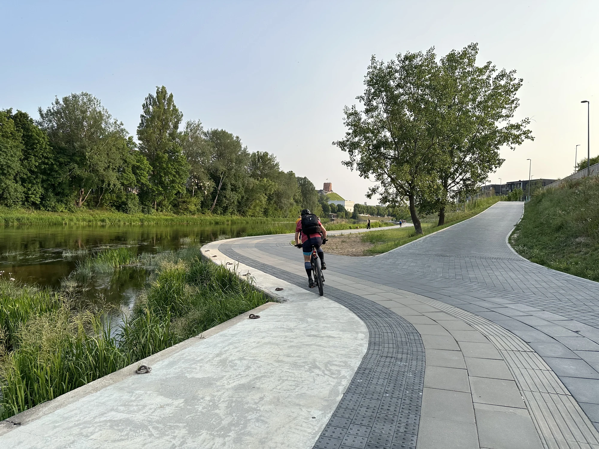 A cyclist riding on a paved riverside path next to the Neris River in Vilnius, with green trees and Gediminas' Tower on a hill in the background.