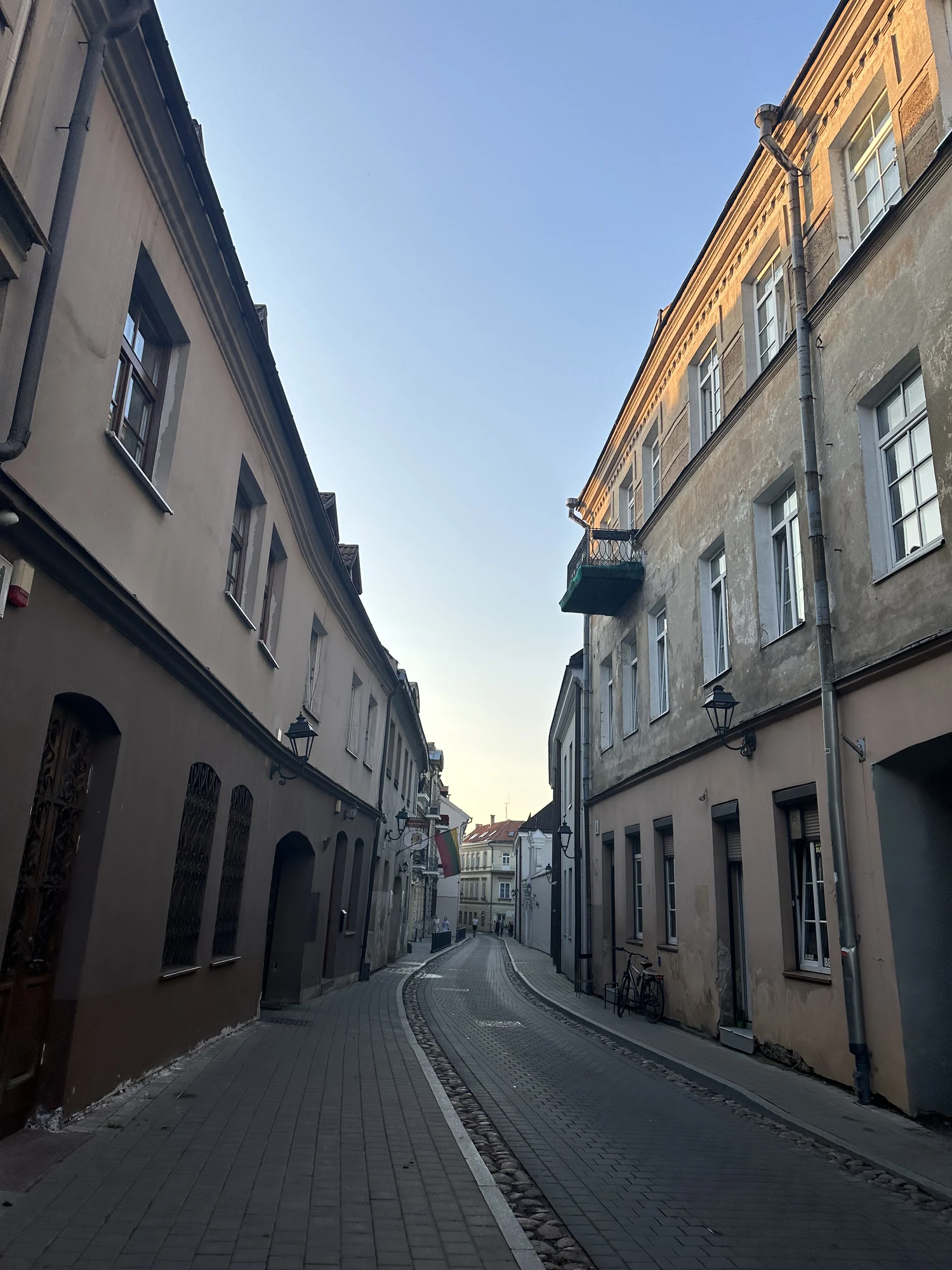 A narrow, curving cobblestone street in Vilnius Old Town, flanked by historic multi-story buildings under a clear blue sky.