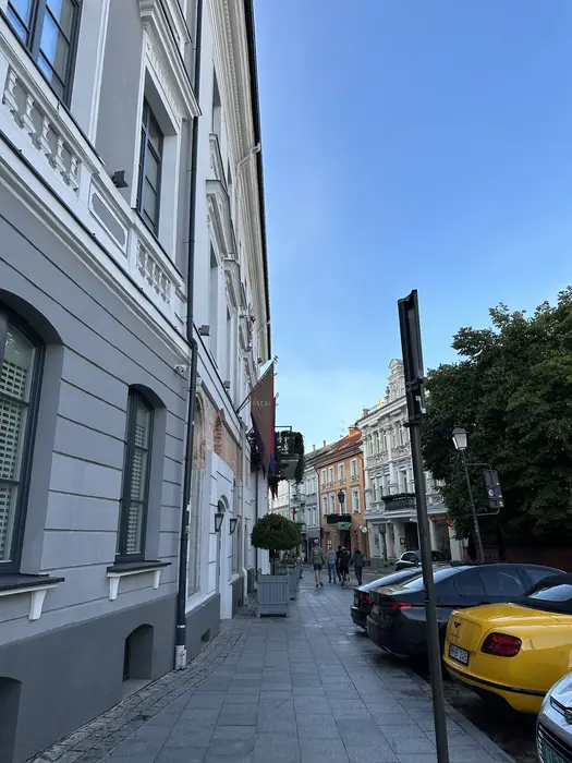 A street-level view of Didžioji Street in Vilnius, featuring the grey facade of Hotel Pacai on the left and parked cars on the right under a clear blue sky.