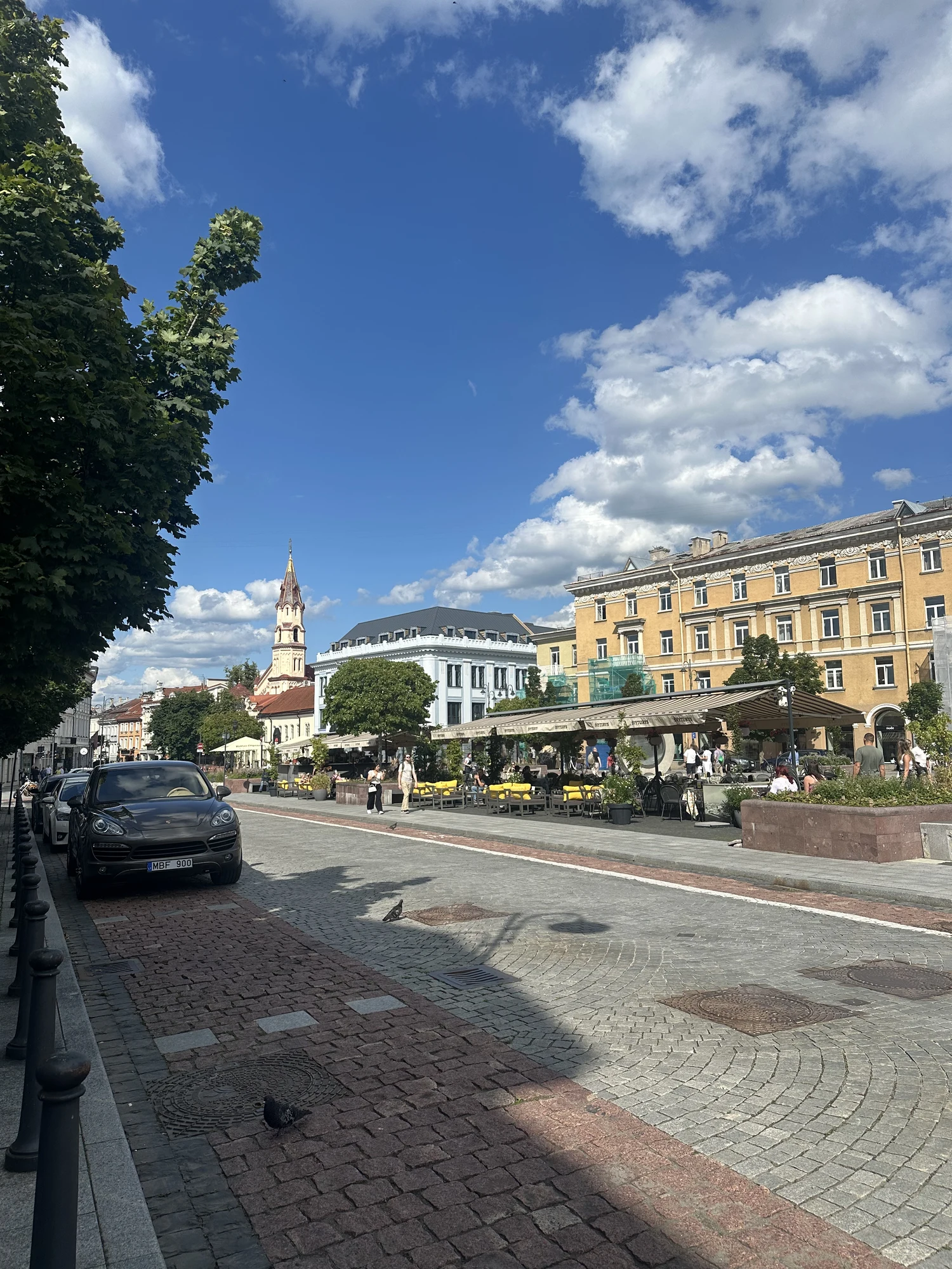 A cobblestone street in Vilnius Old Town featuring outdoor cafes with yellow chairs, historic buildings, a church spire in the background, and a parked car on the left under a blue sky.