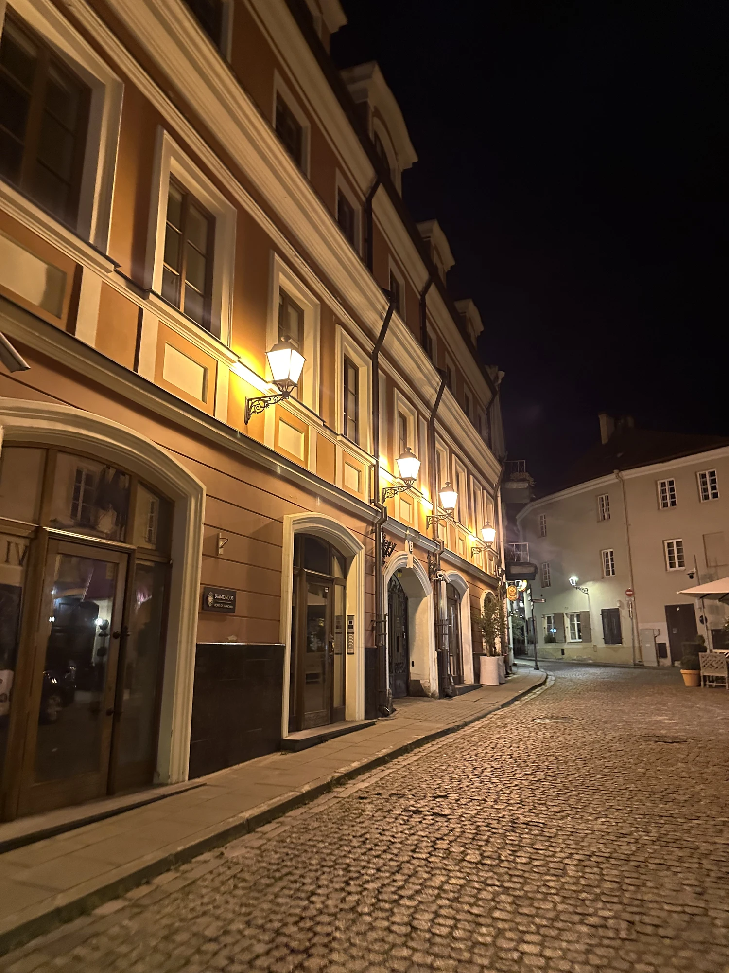 A quiet cobblestone street in Vilnius Old Town at night, lined with historic buildings illuminated by warm wall-mounted lanterns.