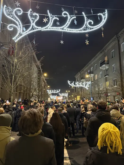A view from behind a dense crowd of people walking down a city street at night, illuminated by bright, star-shaped holiday light arches hanging overhead.