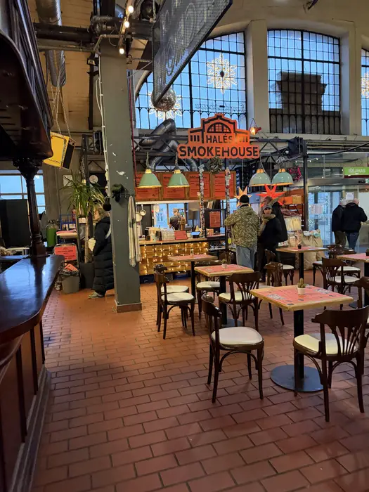An indoor view of Halės Market in Vilnius, showing wooden dining tables and chairs in front of the Halės Smokehouse food stall with customers nearby.