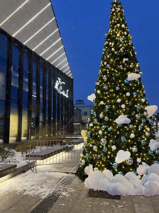 A tall, illuminated Christmas tree decorated with blue and silver baubles and white cloud-shaped ornaments stands next to a modern glass-fronted building with the name 'M.K. Čiurlionis' lit up on its facade at dusk.