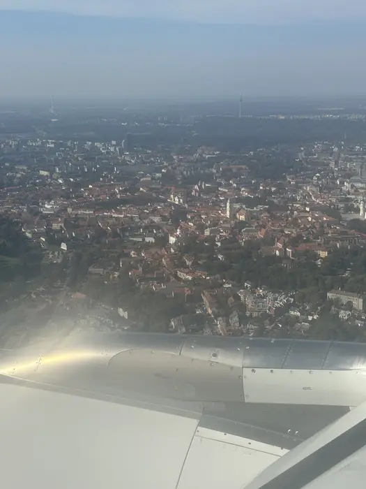 An aerial view of Vilnius Old Town with red-roofed buildings and church spires, seen from an airplane window with the aircraft wing in the foreground.