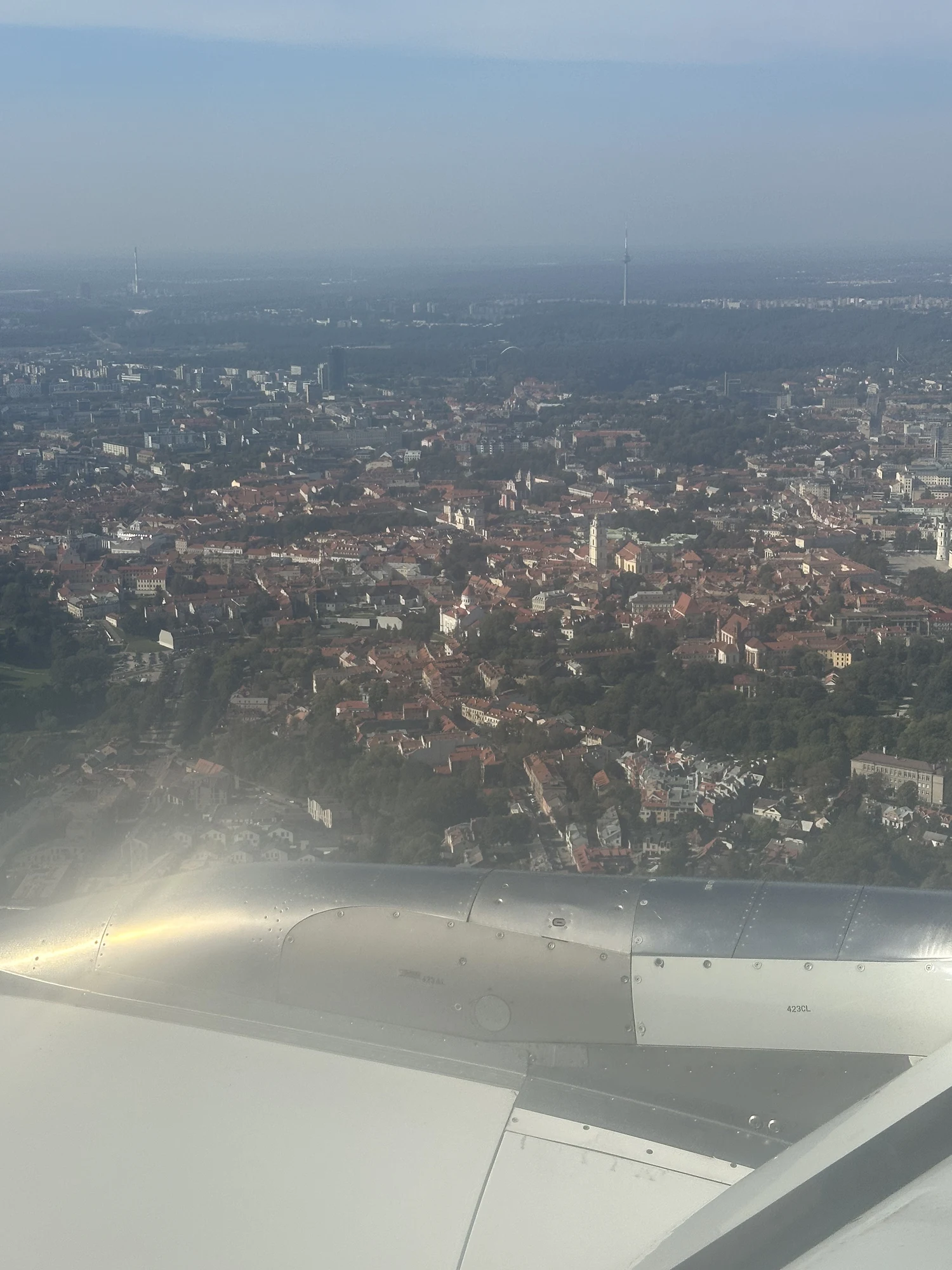 An aerial view of Vilnius Old Town with red-roofed buildings and church spires, seen from an airplane window with the aircraft wing in the foreground.