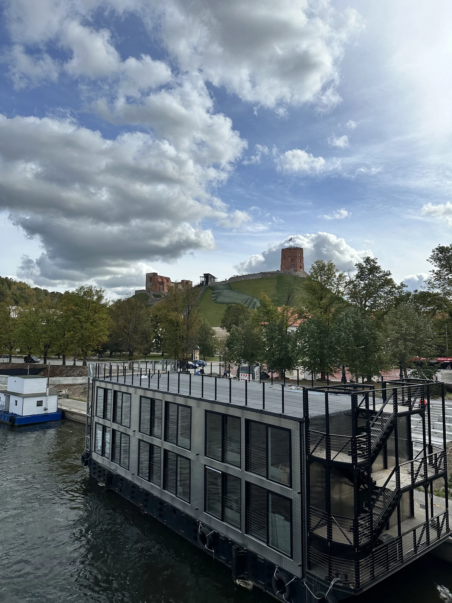 A modern two-story grey houseboat on the Neris River with the historic brick Gediminas' Tower on a grassy hill in the background under a cloudy blue sky.