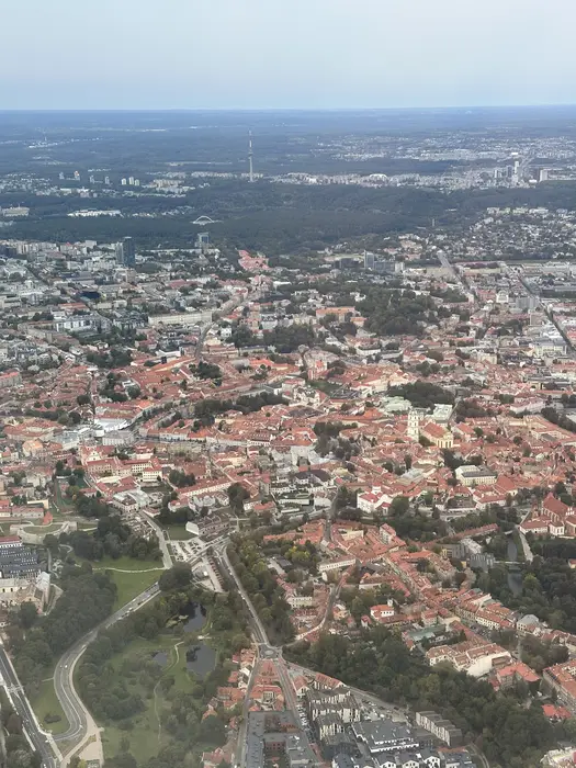 High-angle aerial view of Vilnius Old Town with its dense red-roofed buildings, green parks, and the TV tower visible in the far background under a hazy sky.