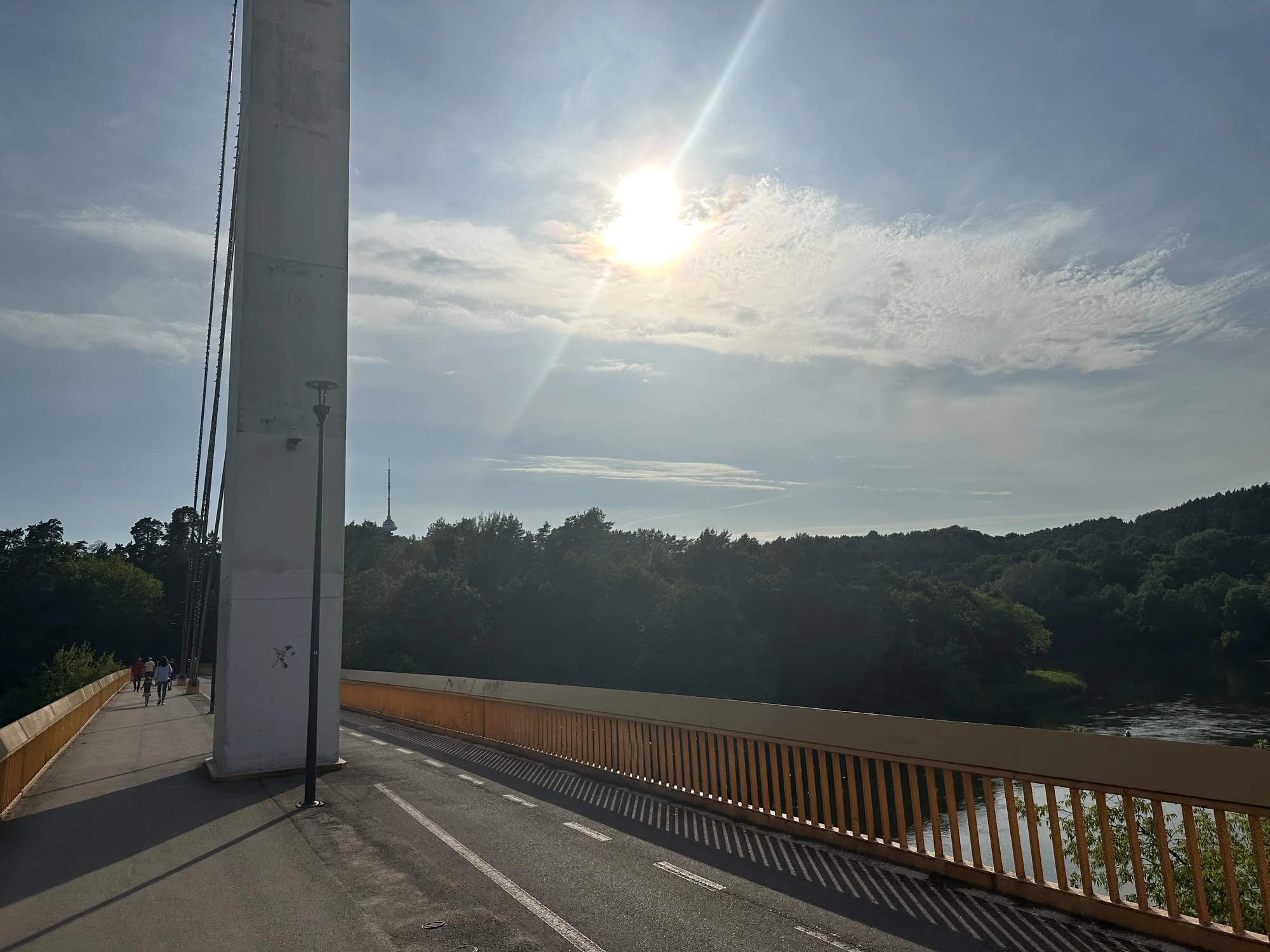 A wide shot of the pedestrian Vingis Park Bridge in Vilnius under a bright sun, with the Neris River and lush green forests in the background.