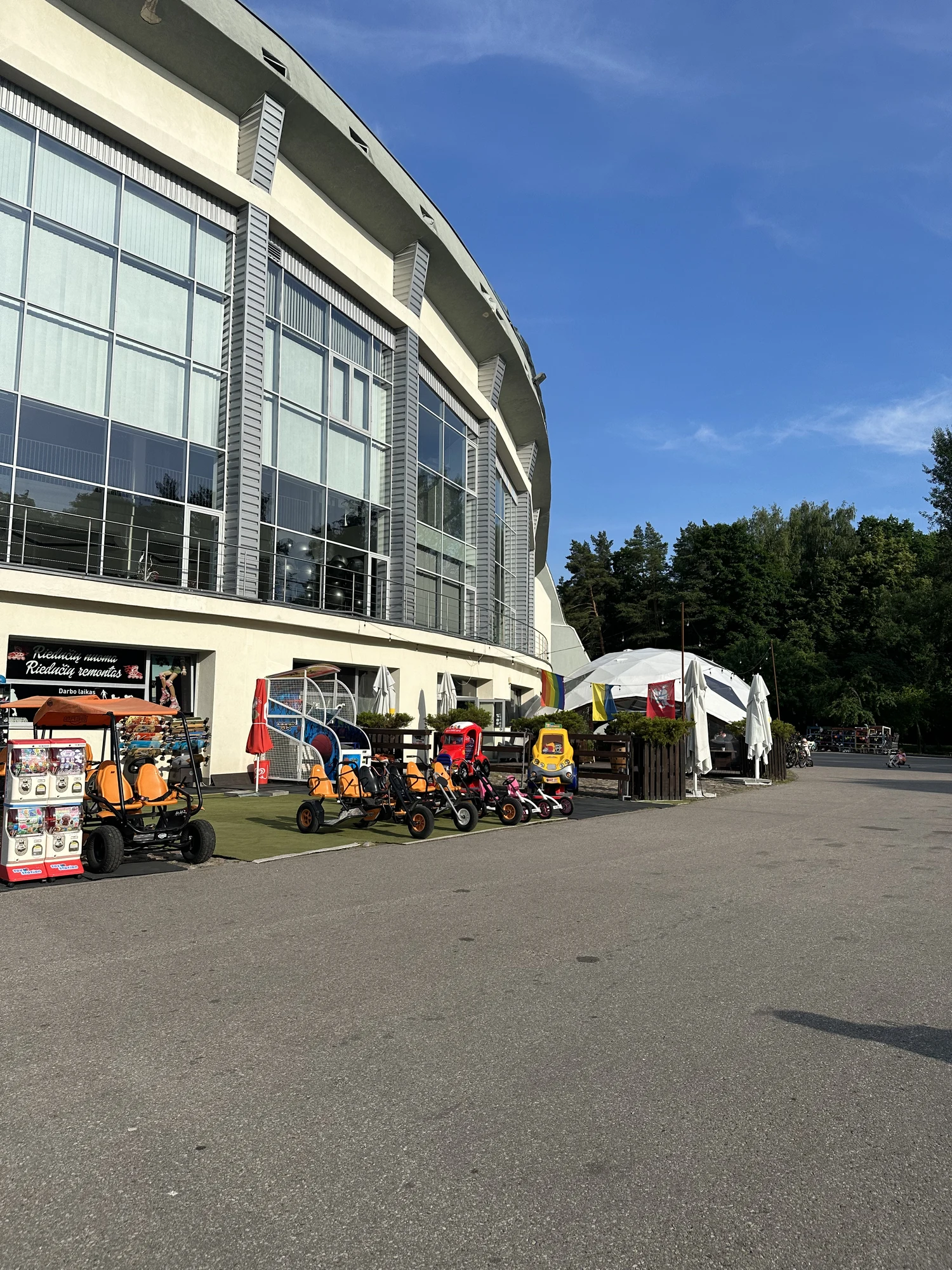 A row of orange and black rental pedal karts parked on the asphalt in front of the curved glass facade of the Vingis Park Stage building under a clear blue sky.