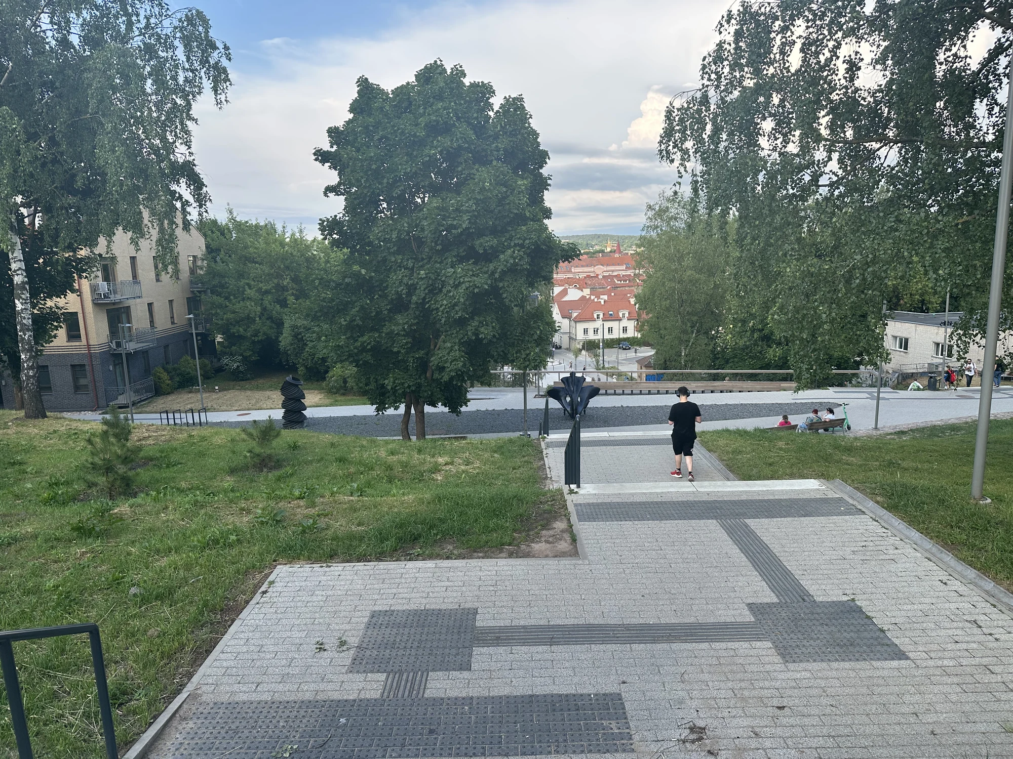 A paved stone pathway with stairs leading down a grassy hill towards trees, sculptures, and red-roofed buildings of Vilnius in the background under a cloudy sky.