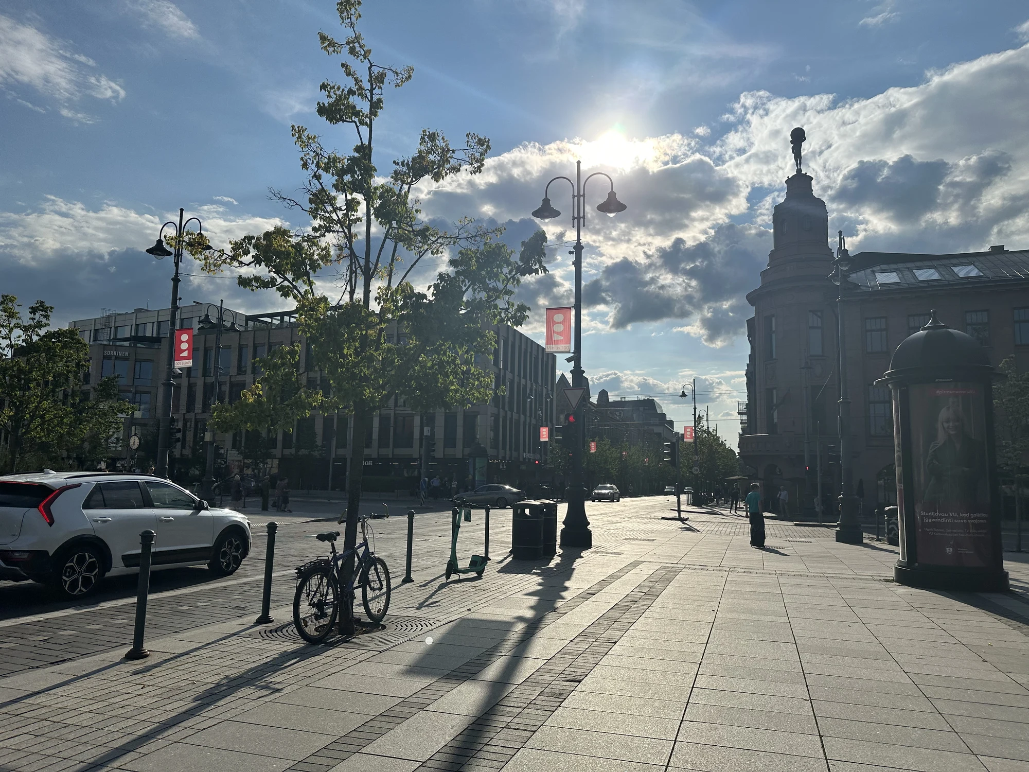 A wide paved sidewalk on Gediminas Avenue in Vilnius under a bright, partly cloudy sky with the sun shining directly ahead. A bicycle is parked next to a young tree, and historic buildings line the street.