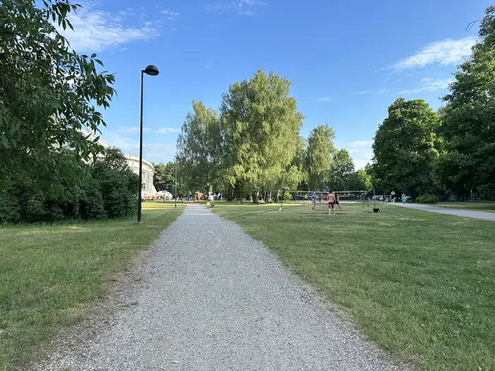 A gravel path cutting through a green lawn in Vingis Park, with people playing volleyball in the background and trees lining the path under a blue sky.