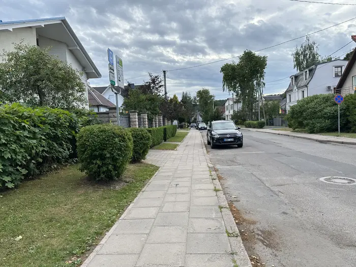 A paved sidewalk runs alongside a quiet street lined with green hedges, houses, and parked cars under an overcast sky.