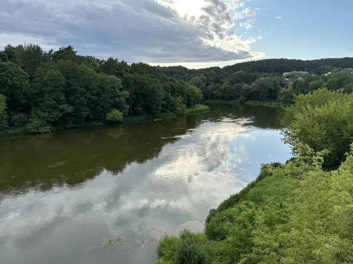 High-angle view of a wide, calm river winding through dense green forests under a cloudy blue sky with reflections on the water surface.