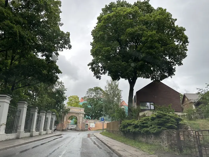 A wet asphalt road in Vilnius leading towards a historic stone archway gate, with a large green tree and a modern brown house on the right.