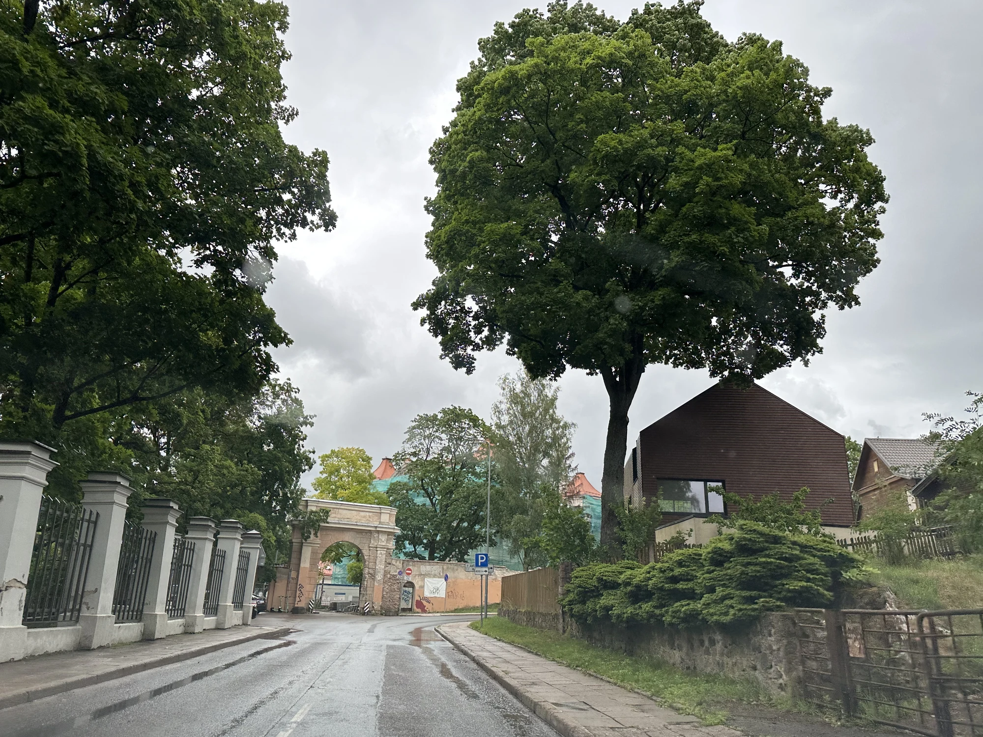 A wet asphalt road in Vilnius leading towards a historic stone archway gate, with a large green tree and a modern brown house on the right.