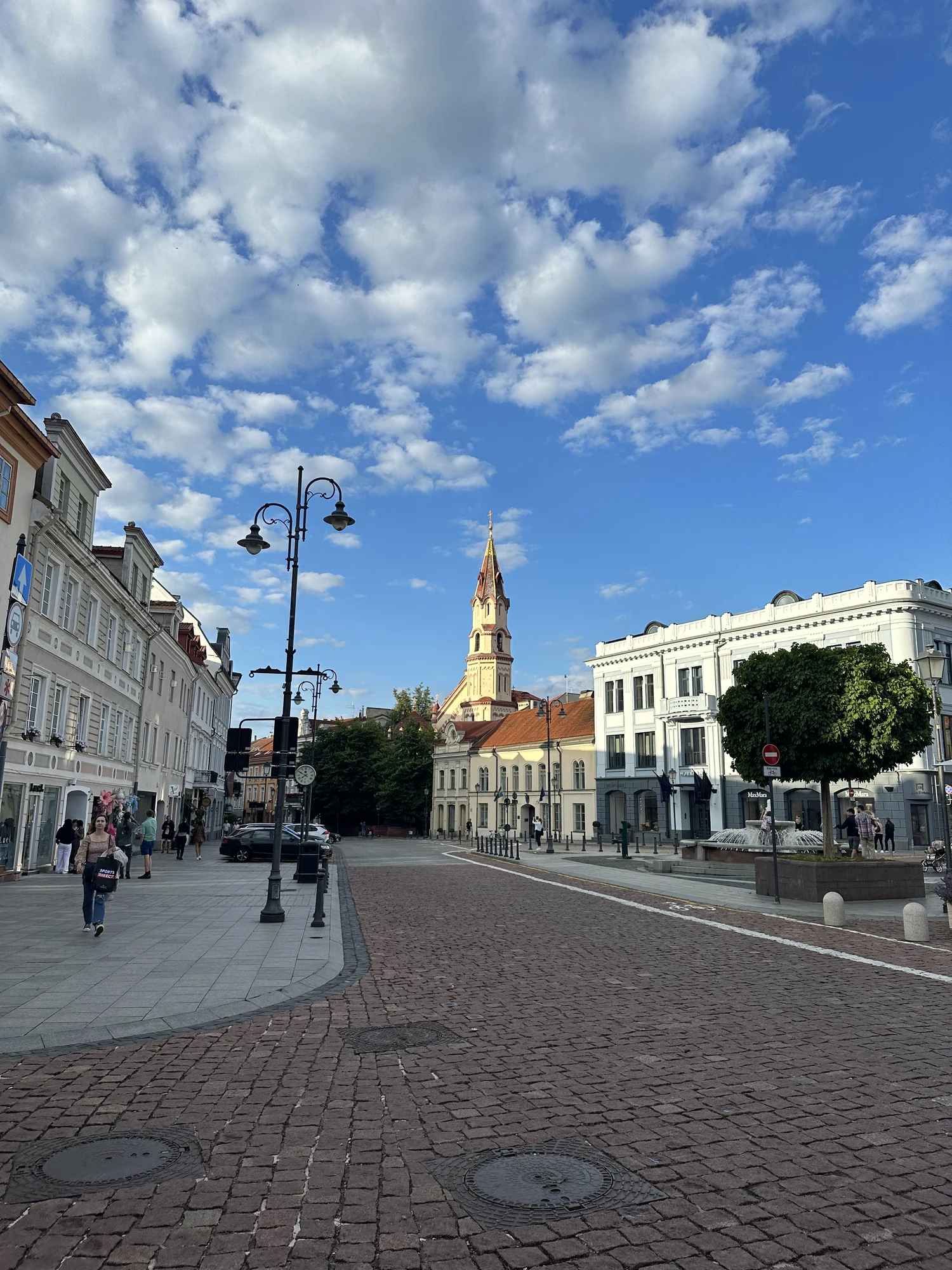 A cobblestone street in Vilnius Old Town with historic buildings on either side, pedestrians on the sidewalk, and a church spire rising in the background under a blue sky with white clouds.