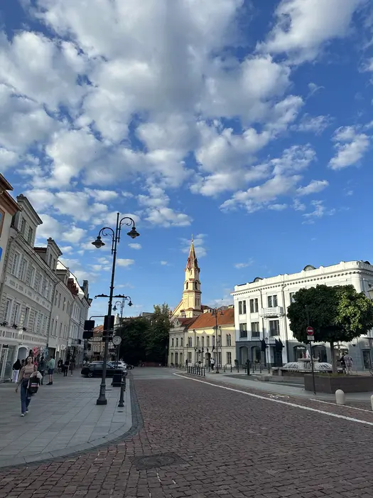 A cobblestone street in Vilnius Old Town under a blue sky with scattered clouds, featuring historic buildings, a streetlamp, and the spire of the Orthodox Church of St. Nicholas in the background.