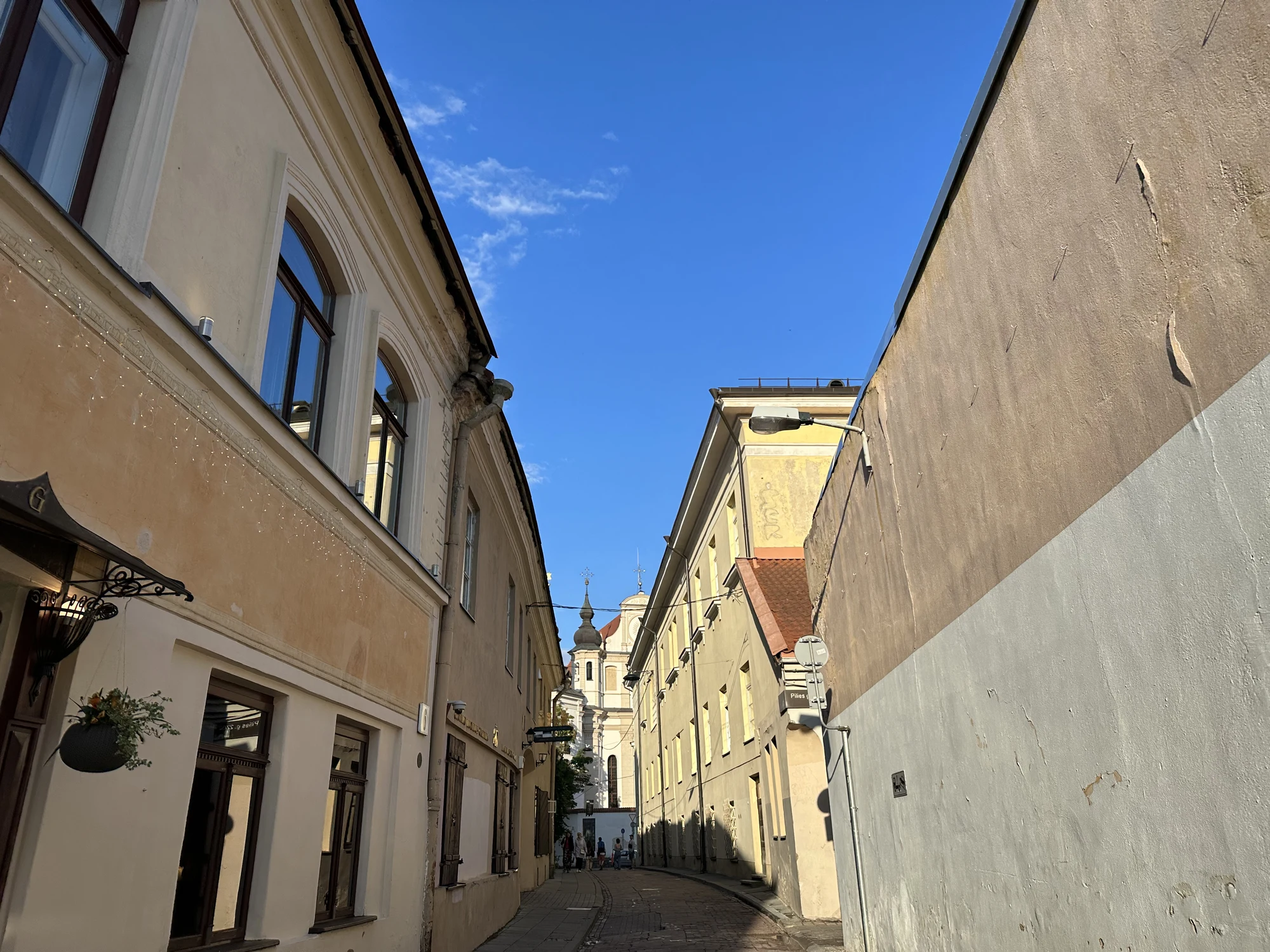 A low-angle shot looking down a narrow cobblestone street in Vilnius Old Town, flanked by historic buildings under a bright blue sky with a church spire visible in the distance.