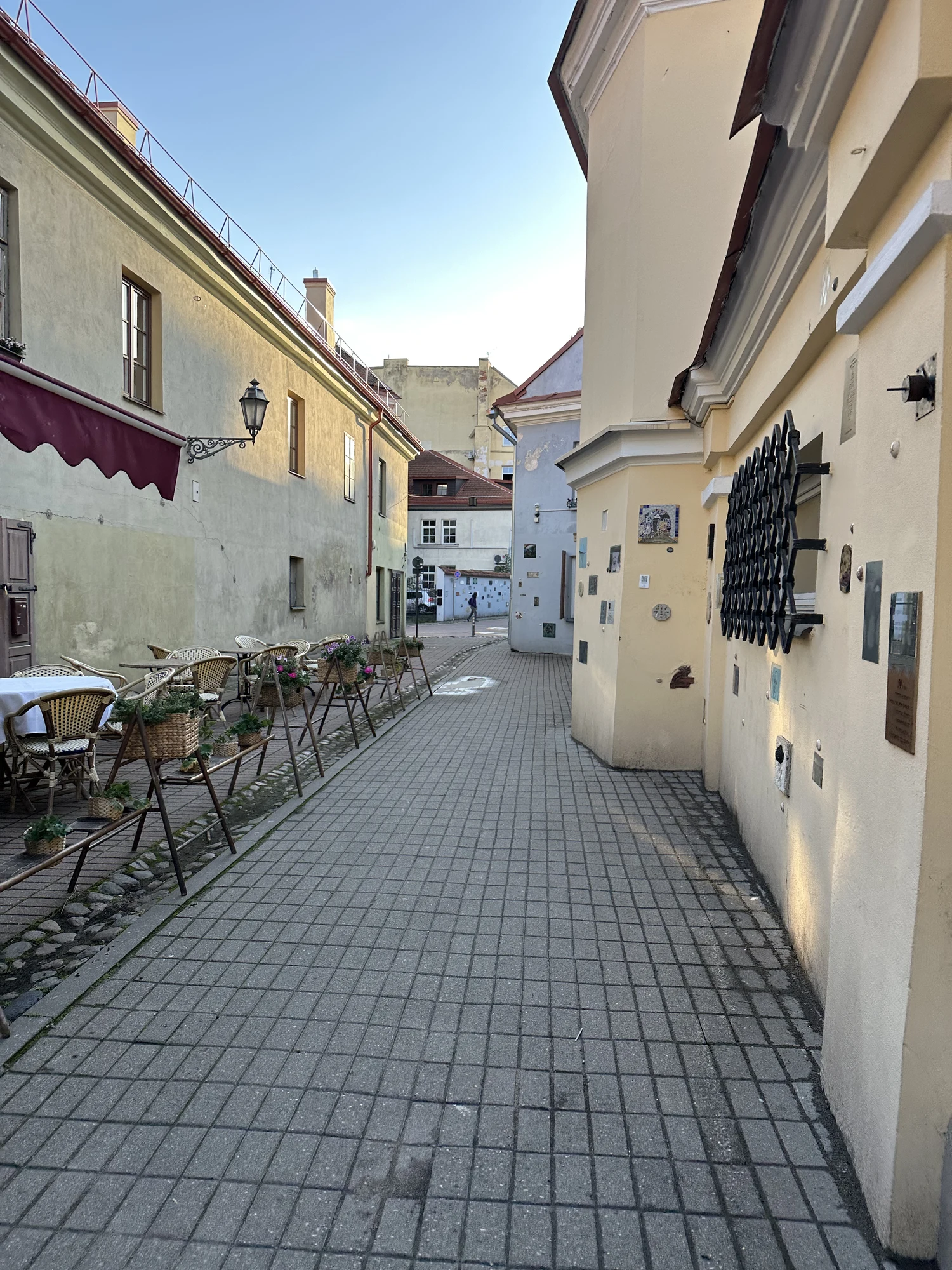 A narrow paved alleyway in Vilnius Old Town lined with historic buildings, outdoor cafe seating on the left, and art plaques on the right wall.