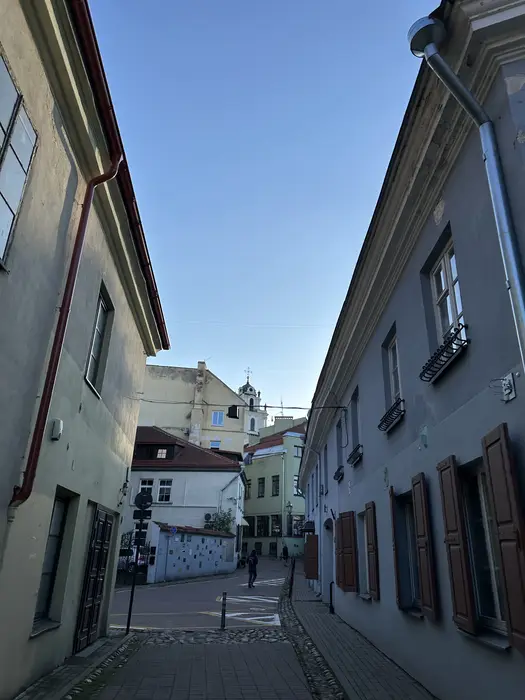 A narrow cobblestone street in Vilnius Old Town lined with historic pastel-colored buildings, leading towards a baroque church tower under a clear blue sky.