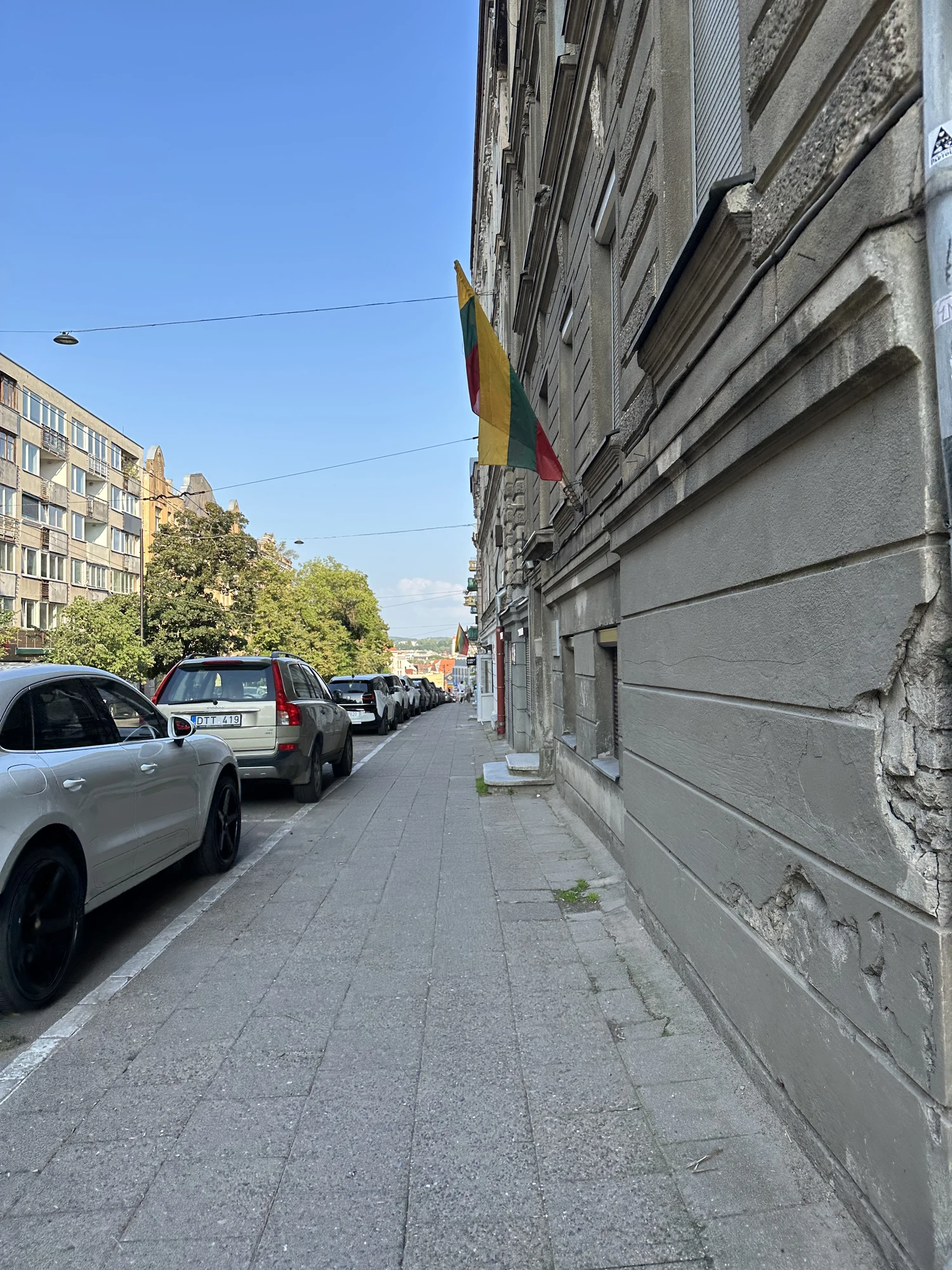 A paved sidewalk in Vilnius lined with parked cars on one side and a grey building facade flying the Lithuanian flag on the other.
