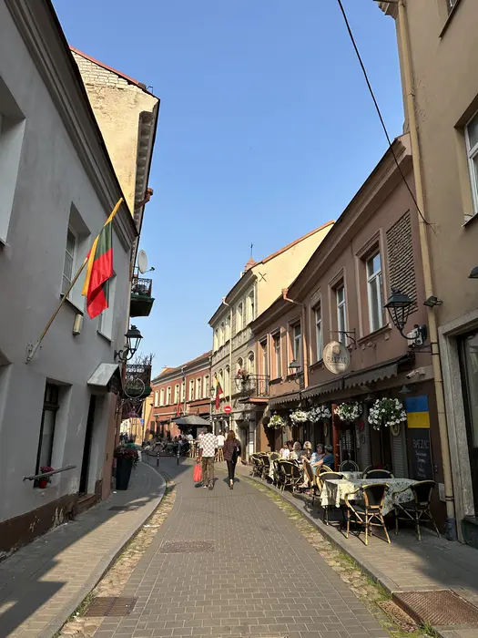 A narrow cobblestone street in Vilnius Old Town lined with historic buildings, outdoor cafe seating, and people walking under a clear blue sky.