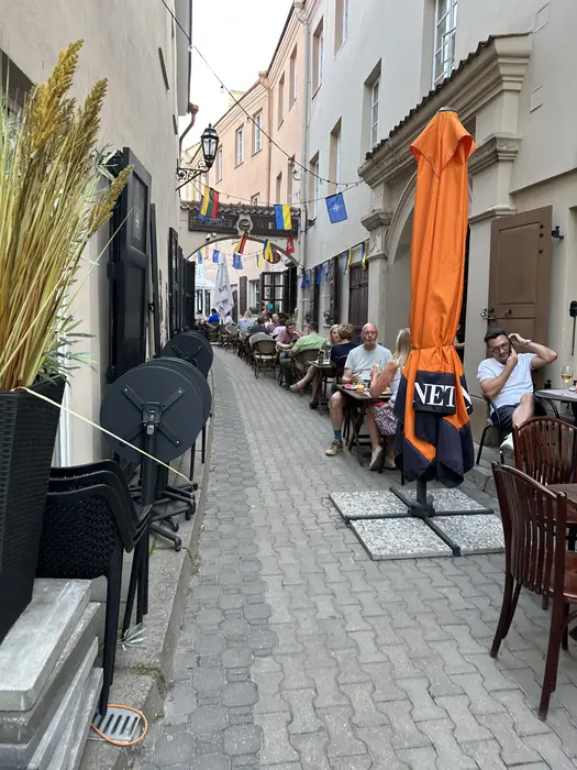 A narrow cobblestone alleyway in Vilnius Old Town lined with outdoor restaurant tables where people are dining under colorful flags.