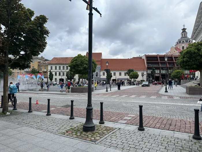 A wide view of Town Hall Square in Vilnius, featuring cobblestone paving, historic buildings, people walking, and the dome of St. Casimir's Church in the background under a cloudy sky.