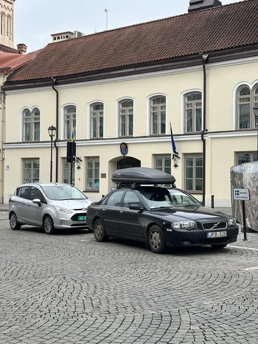 A black Volvo with a roof box and a silver Ford parked on a cobblestone street in front of the pastel-colored Embassy of Sweden building in Vilnius.