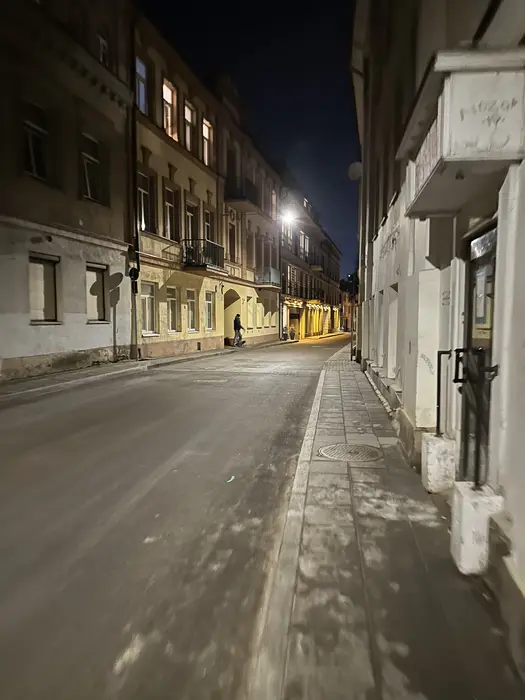 A narrow, paved street in Vilnius Old Town at night, lined with historic multi-story buildings lit by streetlamps, with a lone figure walking in the distance.