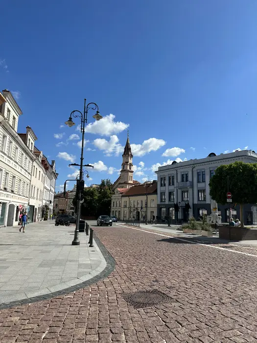 A sunny street view of Didžioji Street in Vilnius Old Town, featuring cobblestone pavement, historic buildings, a decorative street lamp, and a church spire in the background under a blue sky.