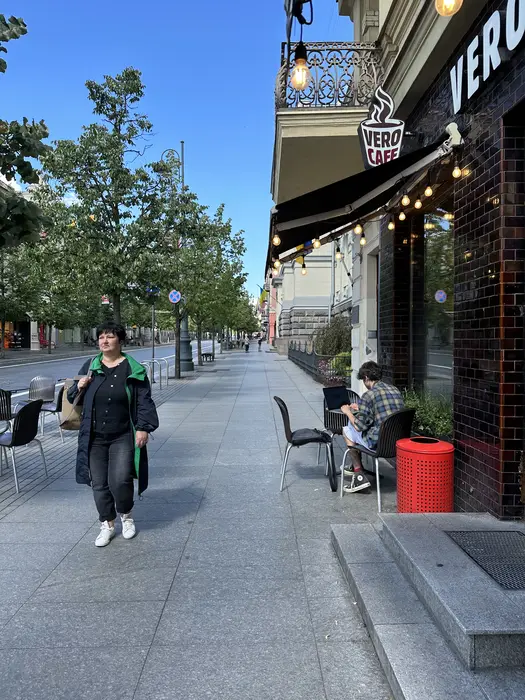 A woman walks down a wide paved sidewalk on Gediminas Avenue in Vilnius, passing a Vero Cafe where a person works on a laptop at an outdoor table.