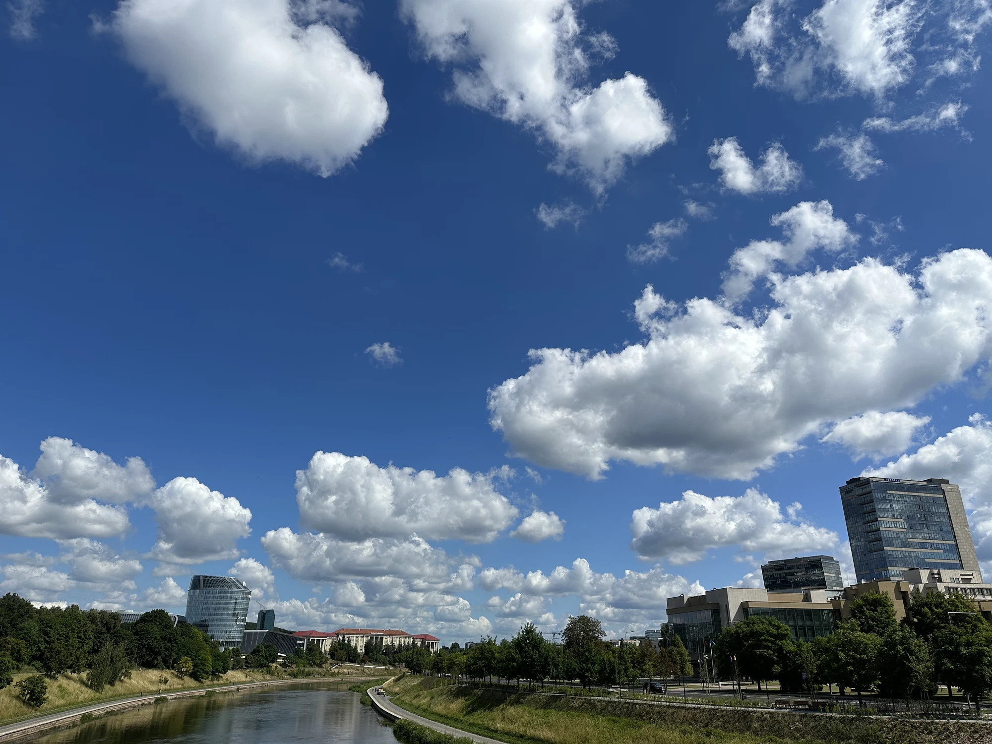 A wide-angle shot of the Neris River in Vilnius, flanked by green banks and modern high-rise buildings under a bright blue sky with scattered white clouds.