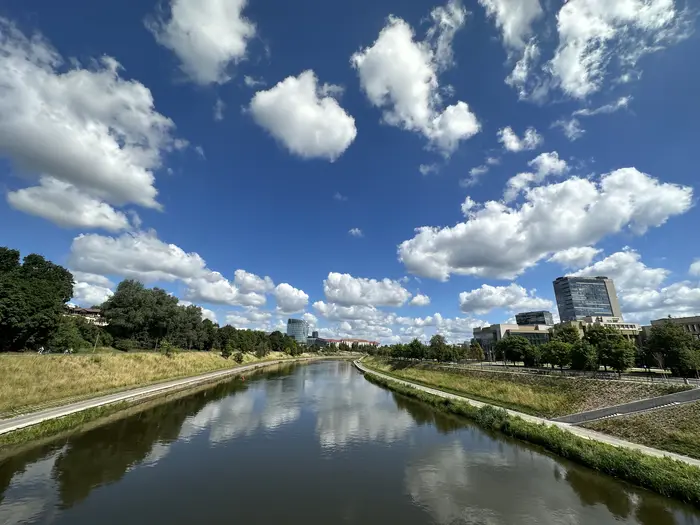 A wide-angle view of the Neris River in Vilnius, Lithuania, reflecting fluffy white clouds under a bright blue sky, with grassy banks and modern high-rises in the background.