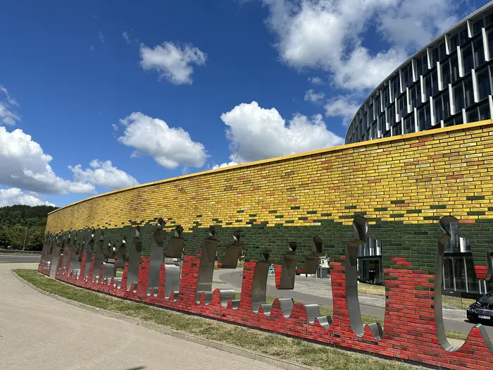 A curved brick wall monument painted in yellow, green, and red with metal cutouts of people holding hands, set against a blue sky with white clouds in Vilnius.