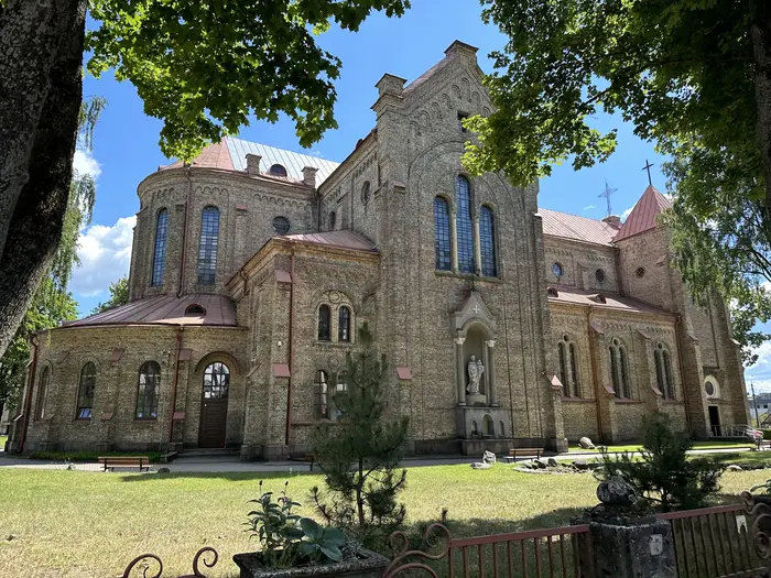 A side view of a large yellow-brick Neo-Romanesque church with arched windows, surrounded by green lawns and framed by lush trees under a bright blue sky.