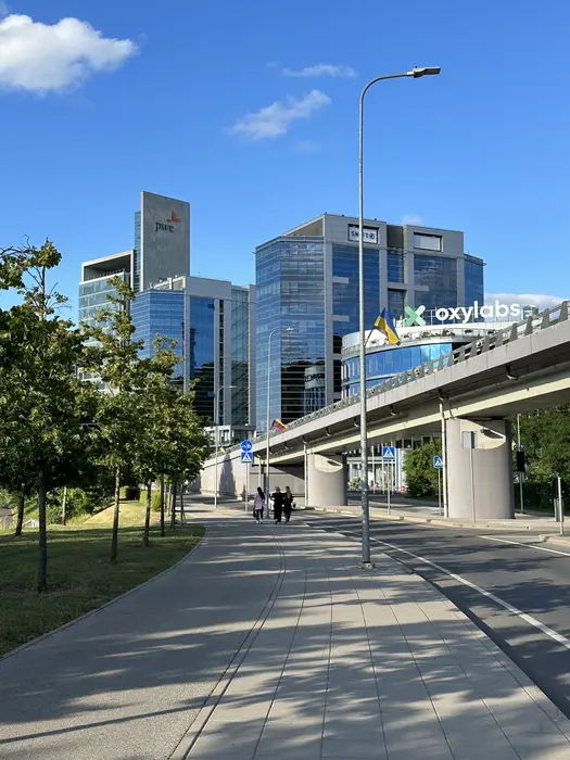 A sunny pedestrian path in Vilnius leading towards modern glass skyscrapers with corporate logos, under a clear blue sky.