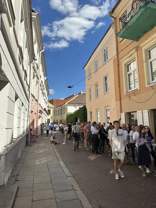 A crowd of people walking and standing along a narrow paved street lined with historic pastel-colored buildings under a blue sky in Vilnius.