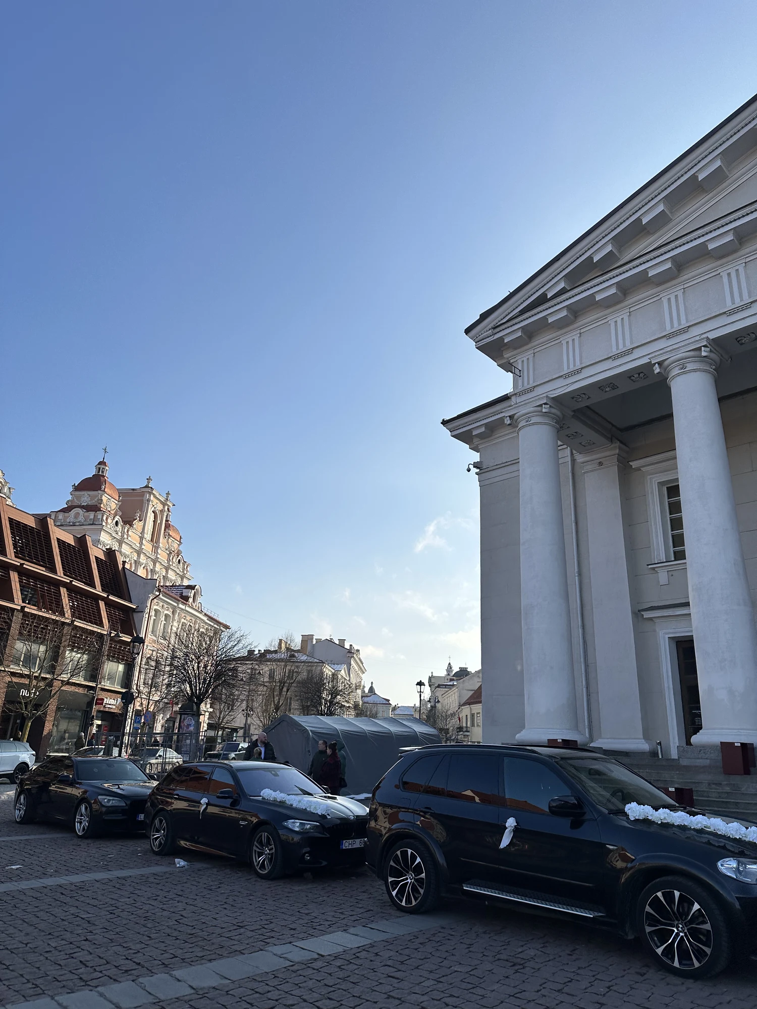Black cars decorated with white flowers parked on the cobblestones in front of the grand columns of the Vilnius Town Hall under a clear blue sky.