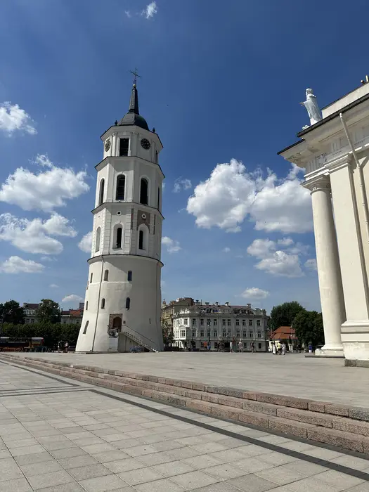 A tall white historic bell tower next to the Vilnius Cathedral on a paved public square under a blue sky with white clouds.