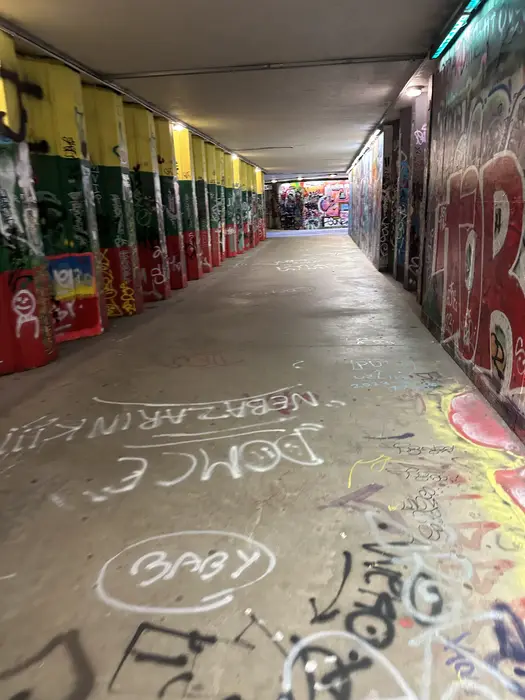 A perspective view inside a concrete pedestrian underpass covered in colorful graffiti, with pillars on the left painted in yellow, green, and red.