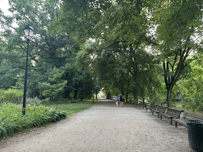 A wide gravel walking path in Bernardine Garden, Vilnius, lined with wooden benches on the right, street lamps, and lush green trees overhead, with two people walking in the distance.