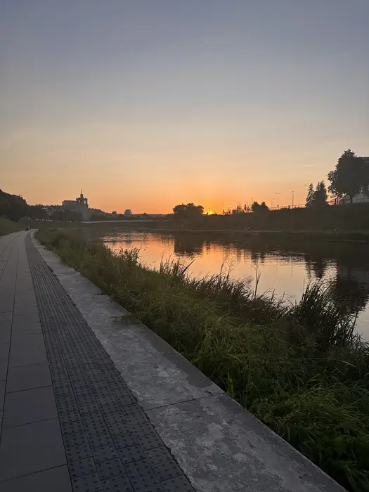 A paved pedestrian path runs along the grassy banks of the Neris River in Vilnius during a vibrant sunset.