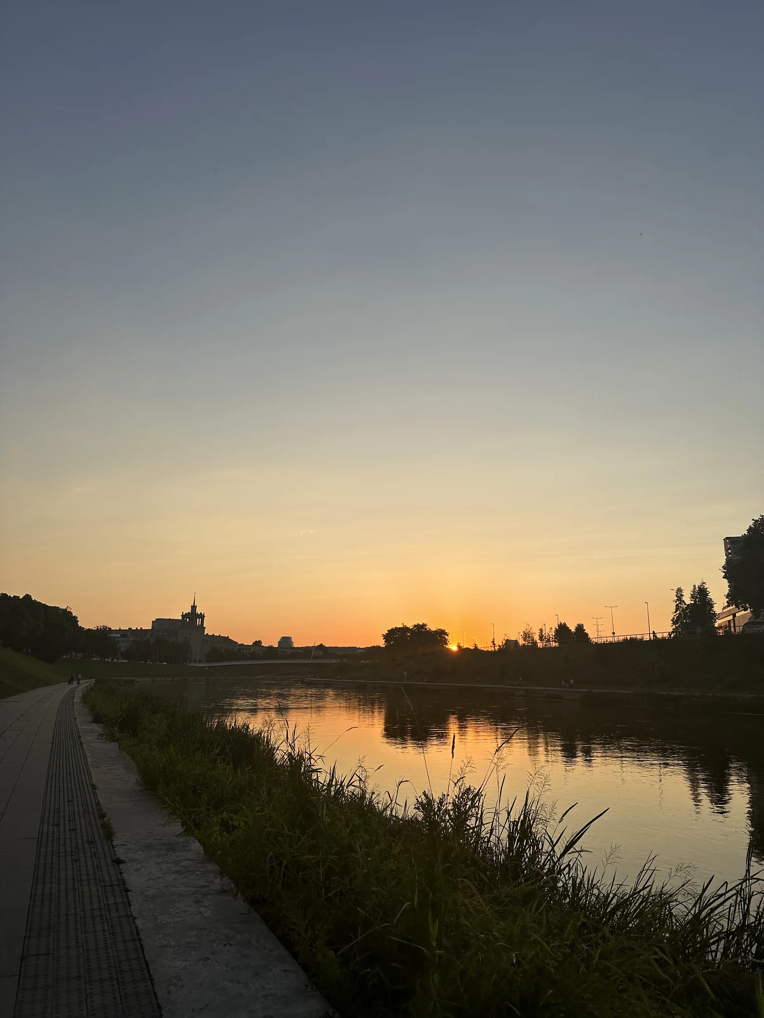 A scenic view of the Neris River in Vilnius during sunset, with a paved walking path on the left and silhouettes of trees and buildings on the horizon.
